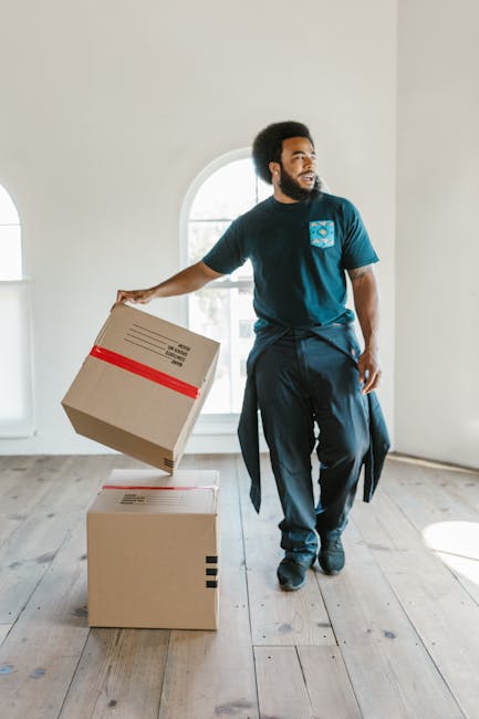 A man with a dark afro hairstyle, beard, and casual attire, including a blue T-shirt and dark trousers, is standing inside a well-lit room with wooden flooring and white walls. He is holding a cardboa