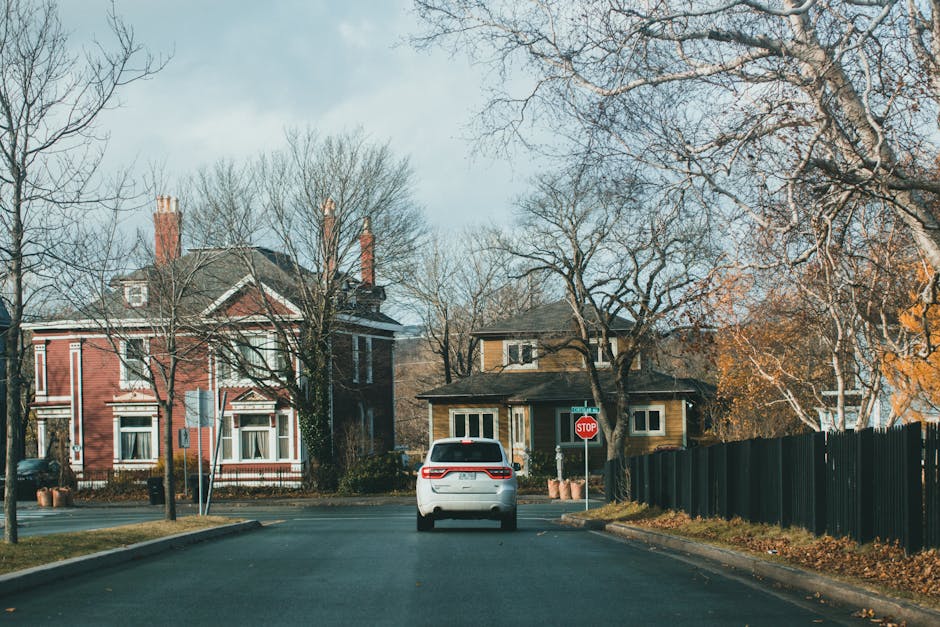 A black and white road sign with the text 'KEEP LEFT' and a downward-pointing arrow located at the edge of a residential street, indicating traffic direction. The street has a paved surface with white
